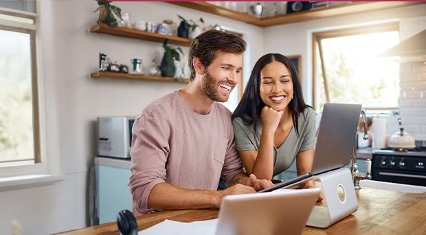 A happy couple sits in their kitchen on their laptop looking at their savings.